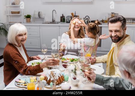 Mädchen, das die Augen der Mutter mit ostereiern bedeckt, während Familie mit Weingläsern toasten, Stockbild Stockfoto