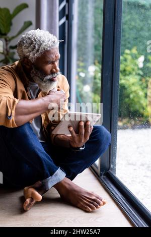 Nachdenklicher afroamerikanischer Mann, der durch das Fenster schaut und auf dem Boden sitzt, mit einem Tablet-pc zu Hause Stockfoto