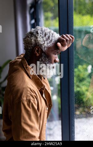 Nachdenklicher bärtiger afroamerikanischer Senior, der sich zu Hause an einem Glasfenster lehnt Stockfoto