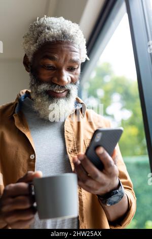 Bärtiger afroamerikanischer Mann mit Smartphone hält Kaffeebecher zu Hause Stockfoto