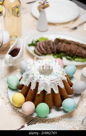 Köstlicher osterkuchen mit bemalten Eiern in der Nähe von Sauce und gebratenem Fleisch auf unscharfem Hintergrund, Stockbild Stockfoto