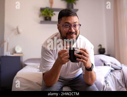 Ein junger Erwachsener lächelt am Morgen auf seinem Bett und trinkt Kaffee Stockfoto
