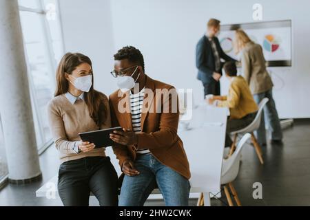 Hübscher junger afroamerikanischer Geschäftsmann und nette Geschäftsfrau, die an einem digitalen Tablet arbeiten, während sie eine Gesichtsschutzmaske tragen Stockfoto