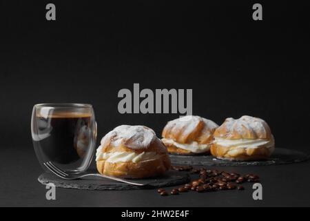 Auf schwarzem Hintergrund, auf schwarzen Tellern liegen Profiteroles mit Pudding, bestreut mit Puderzucker. Eine Tasse Kaffee und Kaffeebohnen komplet Stockfoto