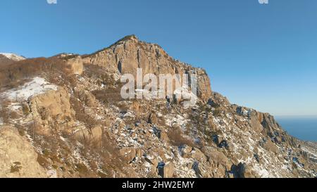 Luftaufnahme der sonnigen Alp Berge im Winter bedeckt von Schnee und kahlen Bäumen auf blauem Himmel Hintergrund. Fliegen Sie an einem sonnigen Tag über steile Klippen. Stockfoto