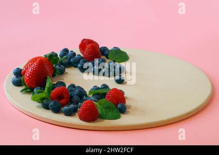 Frische Beeren - Erdbeeren, Himbeeren und Heidelbeeren sowie duftende Minzblätter - werden ordentlich auf einem runden Tablett in Halbmondform gestapelt. Photograp Stockfoto