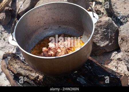 Würzige Brühe mit Rindfleisch kocht in einem Kessel. Zubereitung einer Suppe am offenen Feuer, traditionelles Campingmenü Stockfoto