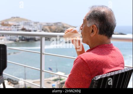 Reifer Mann, der auf einem Balkon mit Blick auf das Meer bei einem Cocktail sitzt Stockfoto