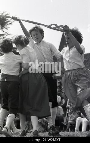 1987, halten sich die Kinder fest an einem Band fest, während sie um den Maibaum, Poppleton, England, Großbritannien, tanzen. Der Maibaum ist ein zeremonieller Volkstanz um einen hohen, mit Blumen und Bändern geschmückten Pol. Er ist eine uralte Tradition, die Jahrhunderte zurückreicht und um Bäume tanzt, um die Ankunft des Frühlings zu feiern. Im Dorfgrün in Poppleton, York, tanzen seit 1945 jedes Jahr Kinder der Grundschule um einen Maibaum herum. Stockfoto