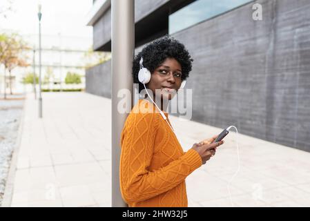 Afro-Frau, die mit einem Smartphone Musik hört, während sie auf dem Bürgersteig steht. Stockfoto