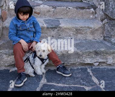 Latino-Junge im Hoodie sitzt mit seinem Pudel auf den Stufen einer argentinischen Altstadt. Horizontal Stockfoto