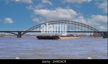 Nijmegen, Niederlande - Februar 27. 2022: Blick über den Fluss waal mit Binnenschiff und Bogenbrücke auf zwei Masten gegen blauen Himmel Stockfoto