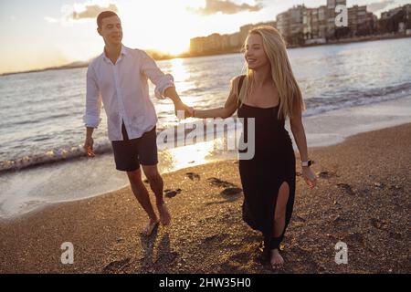 Foto in voller Länge von einem glücklichen Paar, einem Mann und einer Dame, die an der Küste entlang spazieren Stockfoto