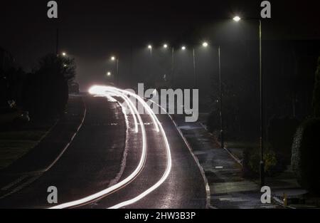 Streifen von Autoscheinwerfern und Heckleuchten, während sie nachts unter der Straßenbeleuchtung, Worcestershire, England, eine Wohnstraße entlang fahren. Stockfoto