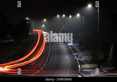 Streifen von Autoscheinwerfern und Heckleuchten, während sie nachts unter der Straßenbeleuchtung, Worcestershire, England, eine Wohnstraße entlang fahren. Stockfoto