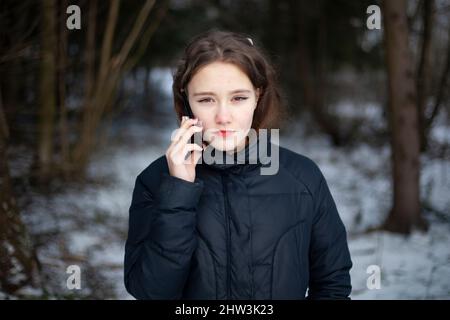 Mädchen telefoniert im Wald. Schulmädchen ist allein im Park im Winter. Frau hält das Smartphone in der Nähe ihres Kopfes. Gehen Sie bei kaltem Wetter. Angespannte Unterhaltung Stockfoto