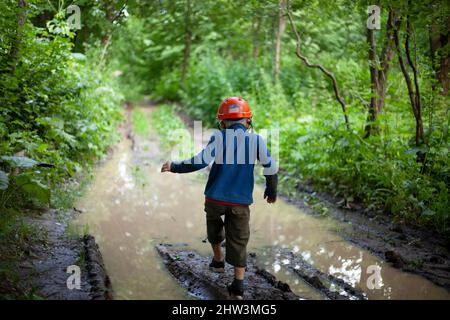 Kind in der Natur. Junge spielt in der Nähe der Pfütze. Vorschulkinder reisen in der Wildnis. Der 6-jährige Junge steht auf einer schlammigen Straße im Wald. Spaziergang im Sommer. Stockfoto