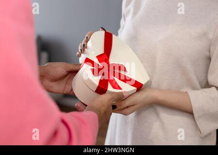 Ein Mann in einem rosa Pullover gibt einem Mädchen in einem weißen Kleid eine weiße Schachtel in Form eines Herzens. Stockfoto