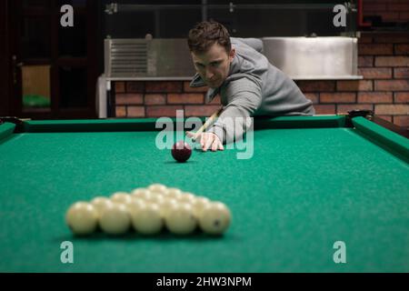 Typ spielt Billard. Der Mensch trifft auf den Ball. Schüler spielt in der Bar Bälle. European hält ein Zeichen, um den Ball zu schlagen. Sportwettbewerb. Konzentrierter, präziser Kick in bil Stockfoto