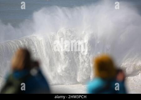 Eine riesige Welle bricht, während zwei weibliche Brillenschauerinnen Ehrfurcht beobachten und fotografieren. Praia do Norte, Nazaré, Portugal. Stockfoto