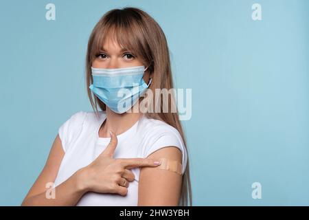 Frau Wurde Gegen Covid-19 Geimpft. Weibchen zeigt geimpften Arm motiviert, gegen Coronavirus zu impfen, um Epidemie zu stoppen. Mädchen in Schutzmaske zeigt Schulter mit Klebeband. Gesundheitswesen Stockfoto