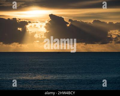 sonnenaufgang am Pazifik, goldenes Sonnenlicht, das durch die Wolken auf das Meer scheint, einsames, silhouetted Segelboot in der Ferne nahe dem Horizont Stockfoto