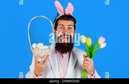 Ostertag. Kaninchenmann in Hasenohren mit Blume. Bärtiger Rüde in Anzug hält Korb mit Eiern. Stockfoto