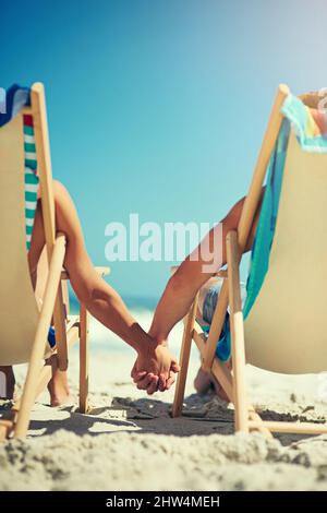 Süchtig nach Liebe. Rückansicht eines jungen Paares, das Hand in Hand auf Liegestühlen am Strand sitzt. Stockfoto