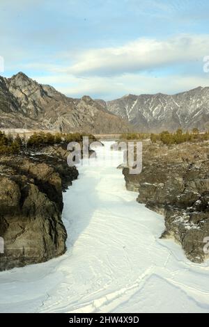 Ein eisbedecktes Flussbett, eingebettet in eine steinerne Schlucht, die durch ein schneebedecktes Tal in den Bergen fließt. Tendelpek Stromschnellen am Katun Fluss, Altai, Stockfoto