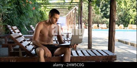 kaukasischer männlicher Geschäftsmann mit Computer sitzt auf einem hölzernen Liegestuhl im Schatten in der Nähe des Pools und arbeitet aus der Ferne. Freiberufler-Konzept im Hotel. cop Stockfoto