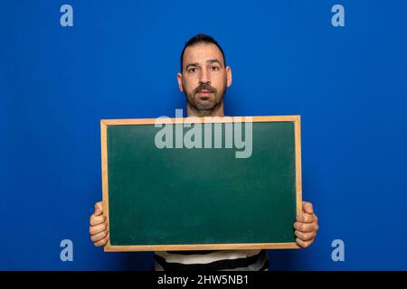 Spanischer bärtiger Mann mit einer Tafel isoliert auf blauem Studiohintergrund. Lern- und Messaging-Konzept. Stockfoto