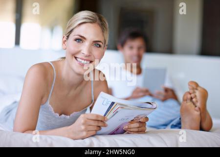 Willkommen am Wochenende. Aufnahme einer glücklichen jungen Frau, die im Bett liegt und ein Magazin liest, während ihr Mann im Hintergrund ein digitales Tablet verwendet. Stockfoto