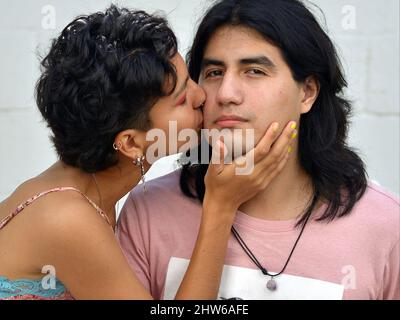 Eine schöne junge Latina-Frau aus Mexiko mit kurzen Haaren küsst einen hübschen jungen Latino-Mann aus Mexiko mit langen Haaren auf der Wange. Stockfoto