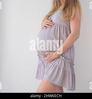 Eine Schwangere in einem blassen lila Nachthemd steht auf weißem Hintergrund und hält ihre Hände auf dem Bauch Stockfoto