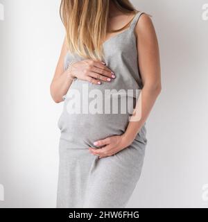 Eine anspruchsvolle Schwangere in einem grauen Kleid steht auf weißem Hintergrund mit den Händen auf dem Bauch, dem Konzept der Weiblichkeit Stockfoto