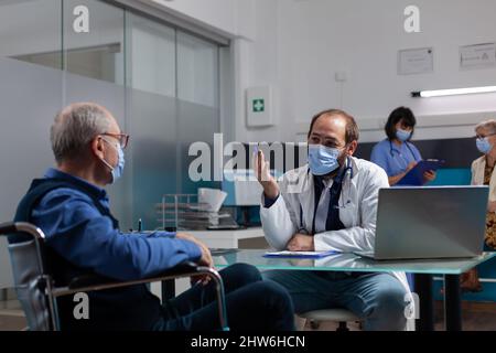 Mann, der während einer Coronavirus-Pandemie im Rollstuhl sitzt, bei einem Arztbesuch. Medic macht Gesundheitsberatung, im Gespräch mit pensionierten Patienten mit körperlicher Behinderung im Büro. Stockfoto