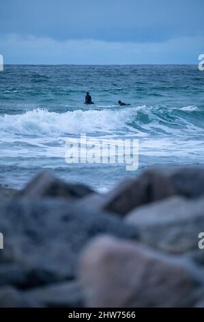 Vertikale Aufnahme eines Ozeans mit zwei Surfern im Wasser an einem bewölkten Tag hinter unbestimmten Felsen Stockfoto