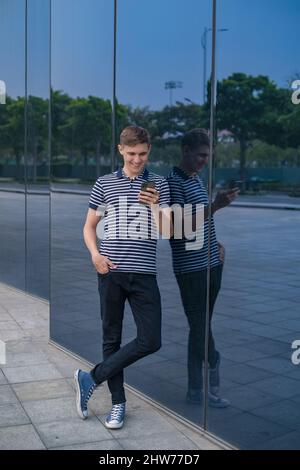 Junger kaukasischer Mann in legerer Kleidung, der in der Nähe des modernen Glasgebäudes steht und sein Telefon anschaut. Personen, die Telefone verwenden. Hochwertige Fotos Stockfoto