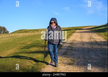 Eine Frau mittleren Alters mit indischer Ethnie, die in niedriger Wintersonne westlich von Ditchling Beacon in East Sussex, England, den South Downs Way entlang geht. Stockfoto