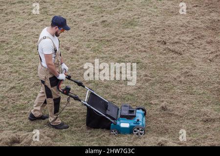 Der Mensch-Gärtner, der Rasen mit einem Vertikutierer erscharft, erscharft den Rasen und entfernt altes Gras Stockfoto