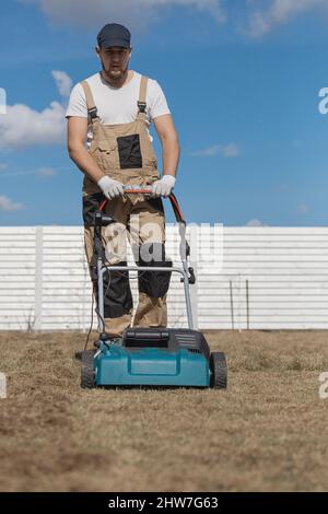 Der Mensch-Gärtner, der Rasen mit einem Vertikutierer erscharft, erscharft den Rasen und entfernt altes Gras Stockfoto