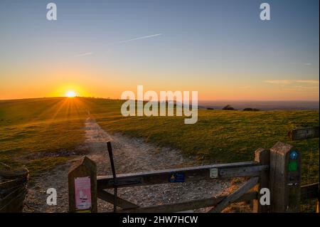 Blick auf eine herrliche untergehende Sonne über Feldern, ein Holztor und den Pfad auf dem South Downs Way westlich von Ditchling Beacon in East Sussex, England. Stockfoto