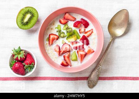 Haferflocken-Haferbrei mit Scheiben Kiwi, Erdbeeren, Mandeln in rosa Schale, Löffel auf weißer Tischdecke Draufsicht Flat Lay Veganes Food-Konzept gesunde Pause Stockfoto
