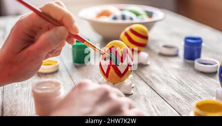 Die Hand der Frau mit einem Pinsel zeichnet ein farblich gefärbtes Muster auf ein Osterei. Vorbereitung auf den hellen Osterurlaub. Stockfoto