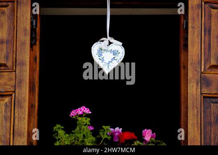 Offenes Holzfenster mit Fensterläden und Blumen mit weißer herzförmiger Dekoration Stockfoto