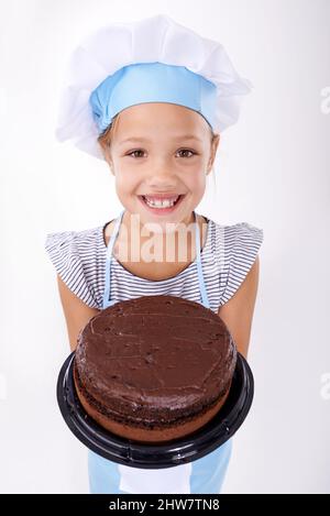 Das ist köstlicher, als es aussieht. Studioaufnahme eines niedlichen jungen Mädchens, das einen Küchenhut trägt und einen Schokoladenkuchen hochhält. Stockfoto