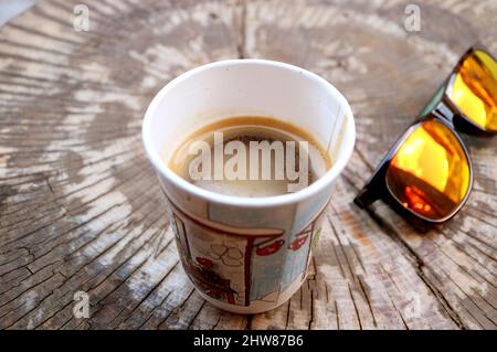 Heißer Kaffee in einer Papiertasse auf einem Baumstumptisch mit Sonnenbrille Stockfoto