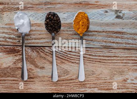 Zutaten aus der Goldenen Paste wie Kokosöl, schwarze Pfefferkörner und Kurkuma-Pulver. Serviert auf Löffel isoliert auf Holzhintergrund mit Kopierraum. Stockfoto
