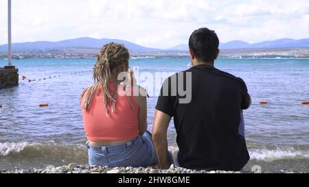 Ein Paar trinkt Wein am Steinstrand. Medien. Junger Mann und junge Frau trinken Wein, sitzen auf einem felsigen Ufer in der Nähe des blauen Meeres. Junges Paar im Urlaub am Meer. Stockfoto