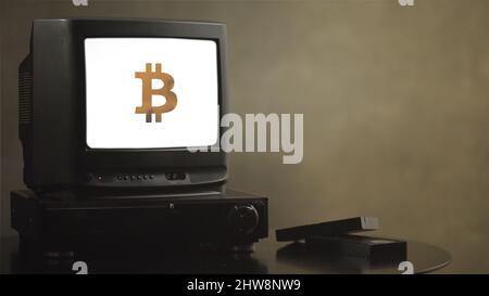 Vintage-Fernseher auf Holztisch mit Bitcoin. Alter Fernseher mit Bitcoin. In der Nähe des Fernsehers befinden sich Filmkassetten und ein Videoplayer. Stockfoto